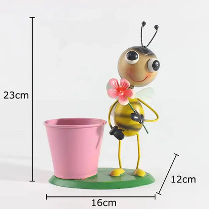 A cheerful yellow and black metal bee figurine holding a pink flower beside a pink bucket on a green base in the ants gardener vase