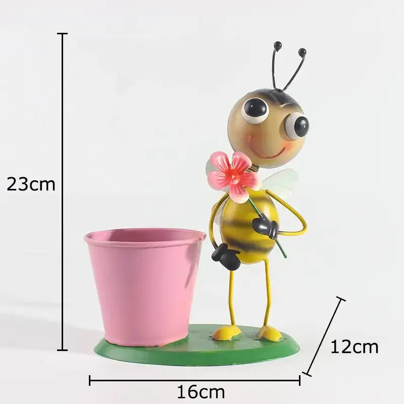 A cheerful yellow and black metal bee figurine holding a pink flower beside a pink bucket on a green base in the ants gardener vase