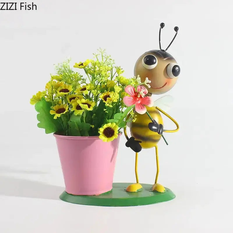 A cheerful yellow and black metal bee figurine holding a pink flower next to a pink pot with artificial sunflowers in the ants gardener vase collection