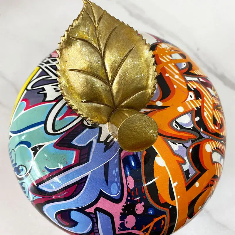 A vibrant glossy pumpkin-shaped decorbites apple figurines art ornament with colorful graffiti and a golden leaf accent