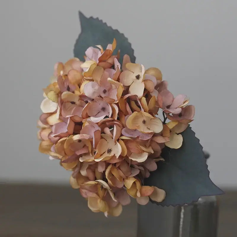 A delicate decorbites artificial hydrangea silk flower arrangement in soft pink peach and cream with dark green paper leaves