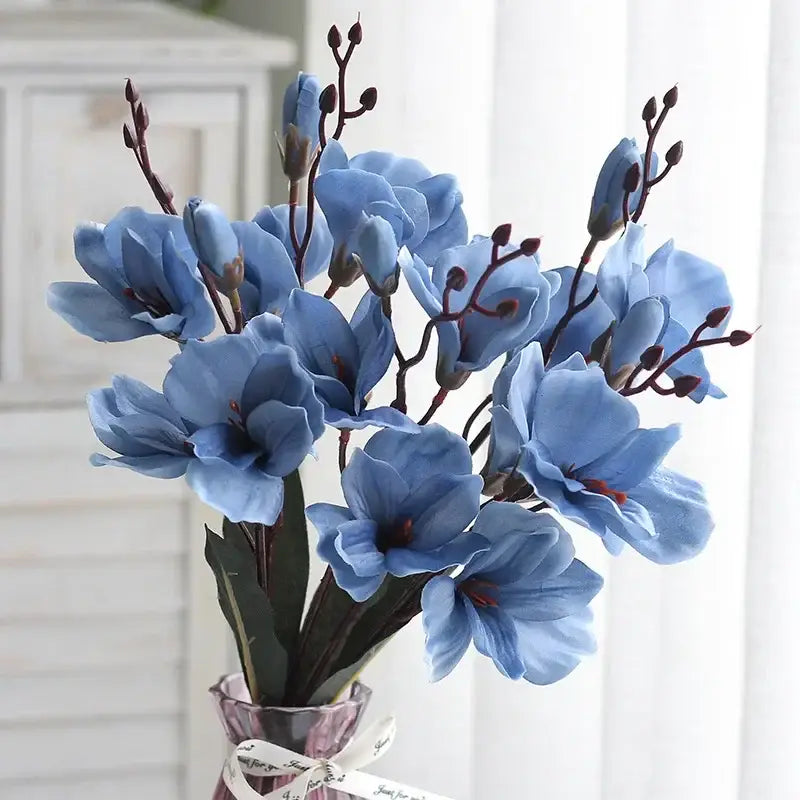 A decorbites artificial orchid magnolia bouquet of light blue flowers with dark stems in a glass vase with white ribbon