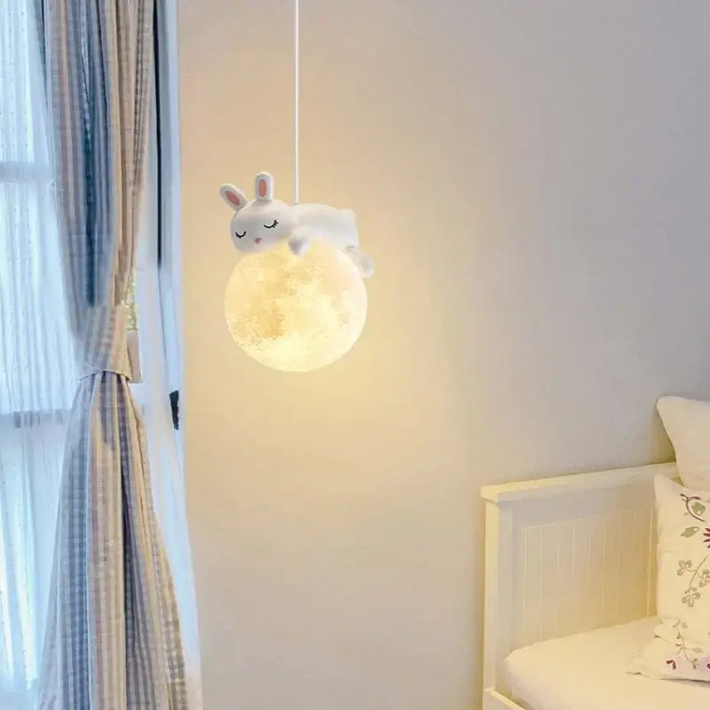 A soft white plush rabbit with closed eyes rests on a glowing textured moon pendant light in the decorbites astronaut chandelier