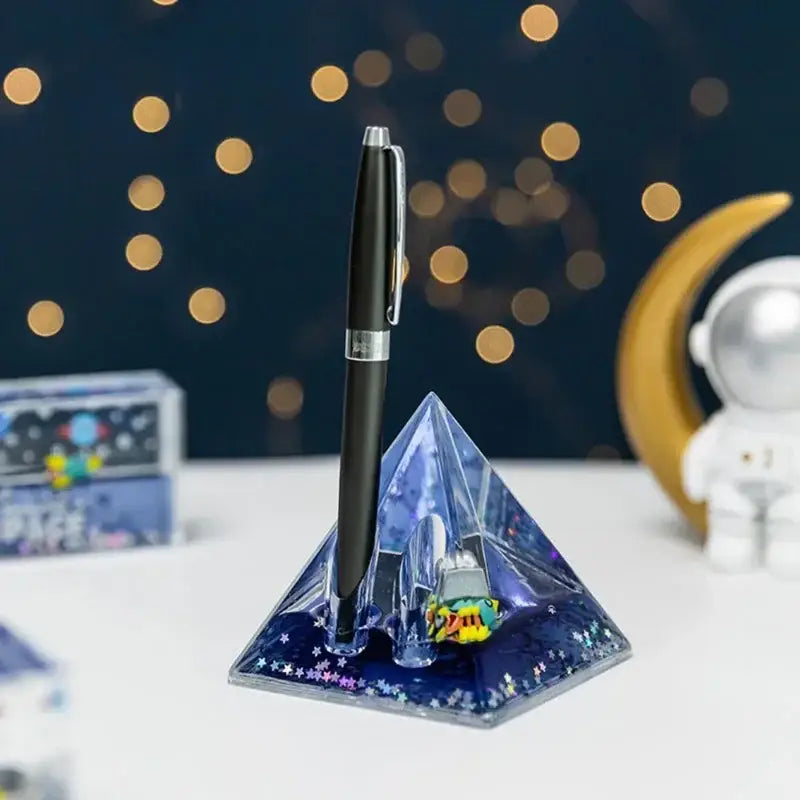 A sleek black pen with silver clip rests in a starry blue pyramid desk holder featuring decorbites astronaut drift design
