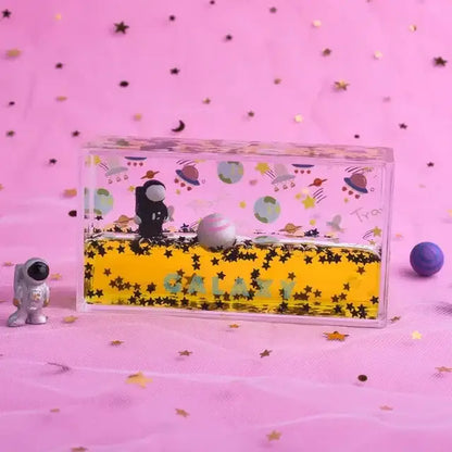 Clear rectangular galaxy-themed liquid-filled keepsake box with black astronaut figurine floating in yellow liquid decorbites astronaut drift