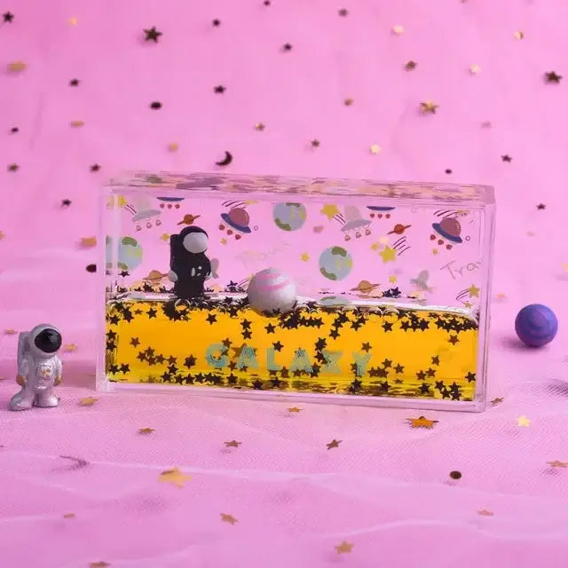 Clear rectangular galaxy-themed liquid-filled keepsake box with black astronaut figurine floating in yellow liquid decorbites astronaut drift