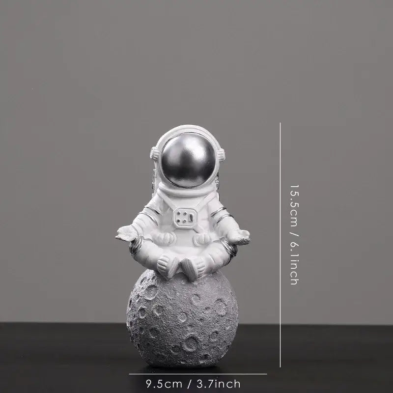 White astronaut figurine with metallic silver helmet seated in meditation pose on textured gray moon surface decorbites astronaut