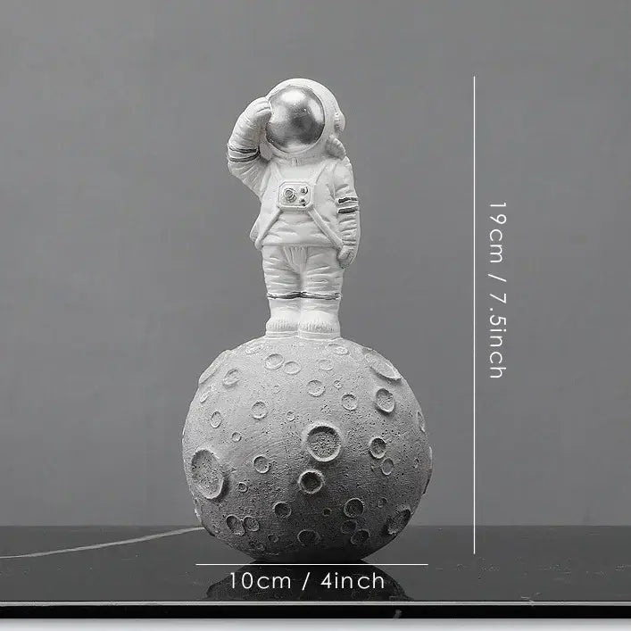 A white decorbites astronaut figurine standing on a textured gray moon surface with one hand raised to helmet