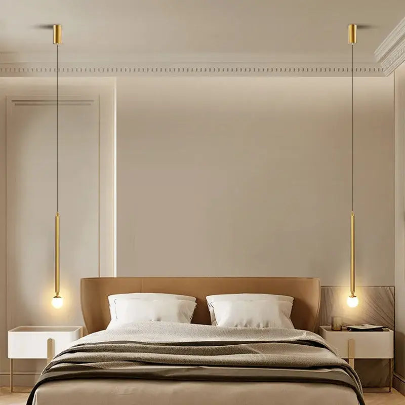 Two slender gold-finished decorbites black gold hanging pendant lights with cylindrical shades above a bed