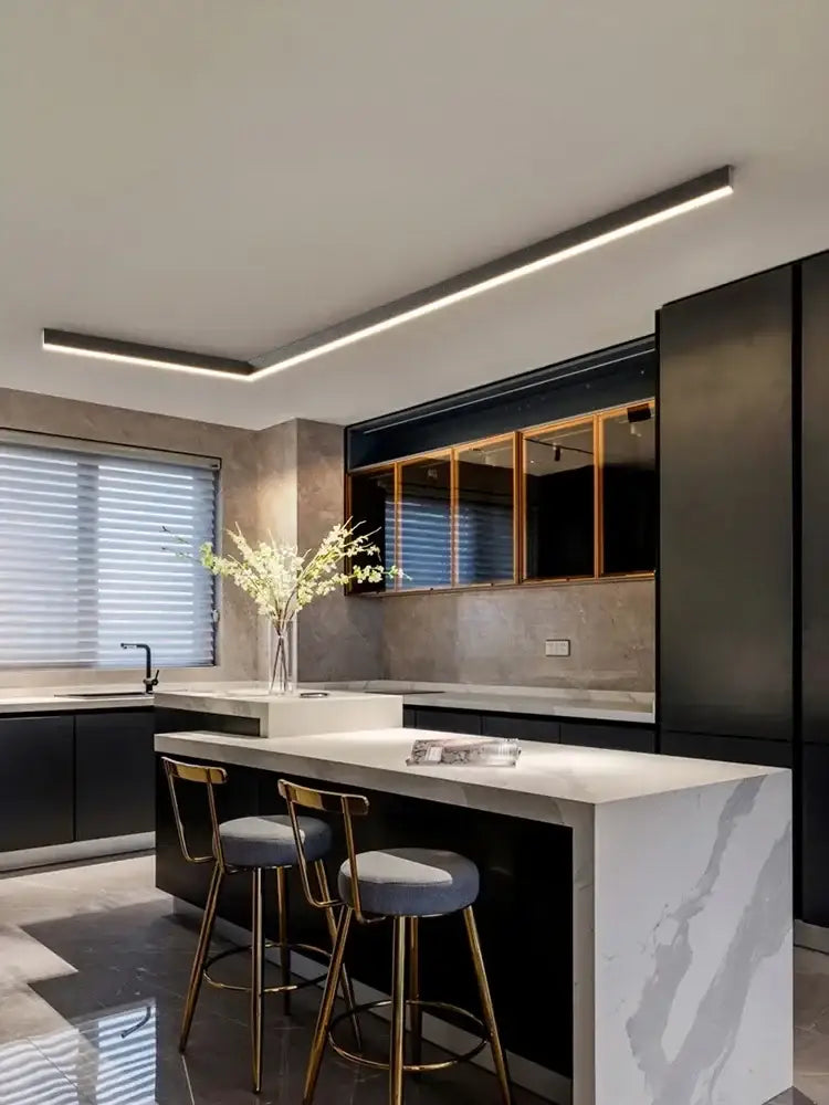 Modern black and white marble kitchen island with gold-toned bar stools under a decorbites black linear led ceiling lamp