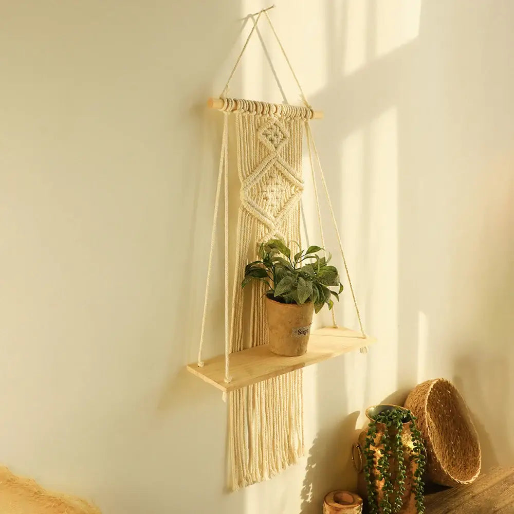 Cream-colored macrame hanging shelf with wooden dowel and fringe holding potted plant for boho home decor wall shelf