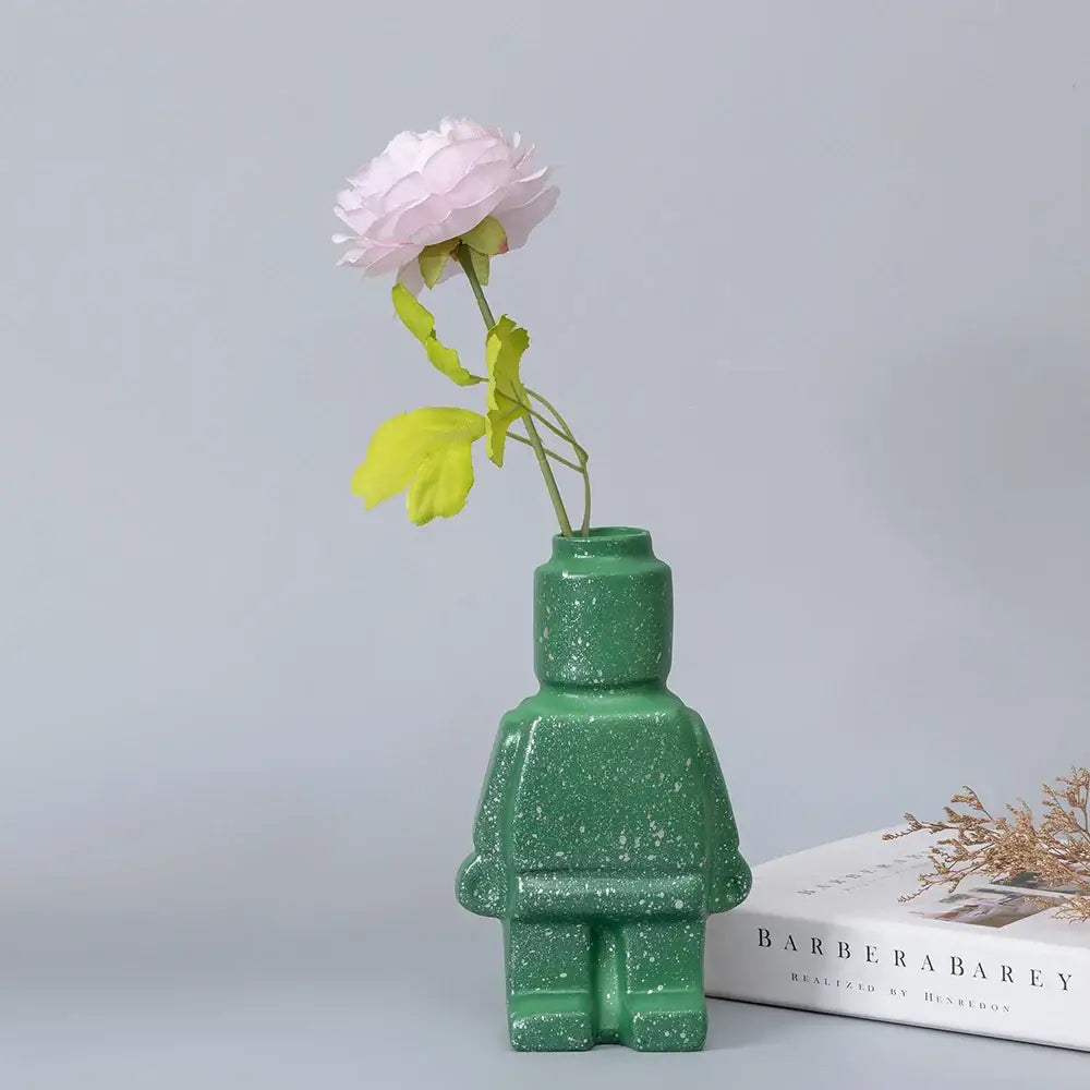 Green glittery ceramic robot vase in nordic style holding a single pink flower for home decor