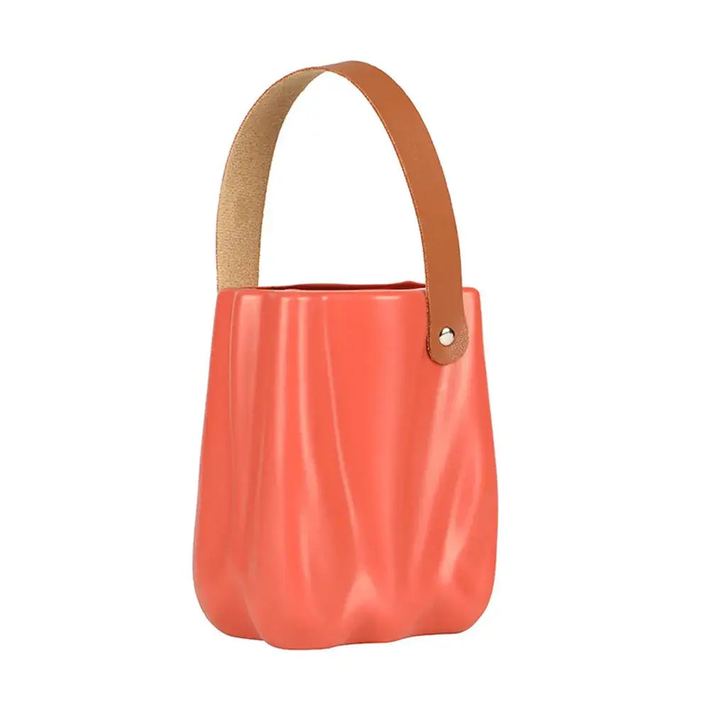 Coral-colored glossy bucket bag with tan leather handle and silver hardware alongside a chic ceramic vase for home decor
