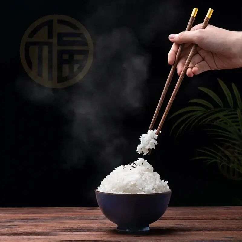 Dark brown chicken-wing wood chopsticks set with gold tips lifting steaming rice from deep blue bowl decorbites