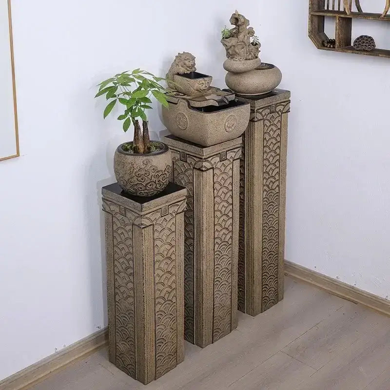 Three beige intricately carved stone stake display columns with wave patterns supporting planters and a small fountain