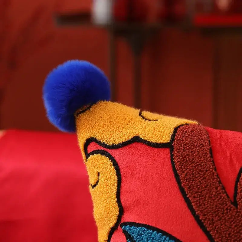 A plush red and yellow dragon embroidered cushion cover with black outlines and a bright blue fuzzy pom-pom topper