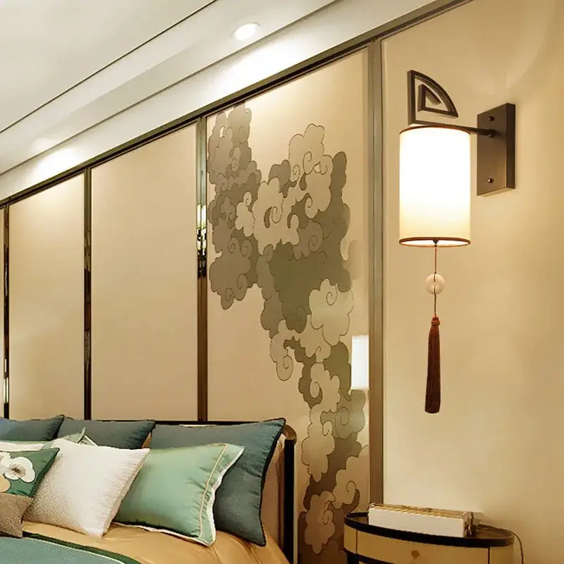 A modern cloth wall sconce with off-white cylindrical shade, dark metal bracket, and decorative tassel for living rooms