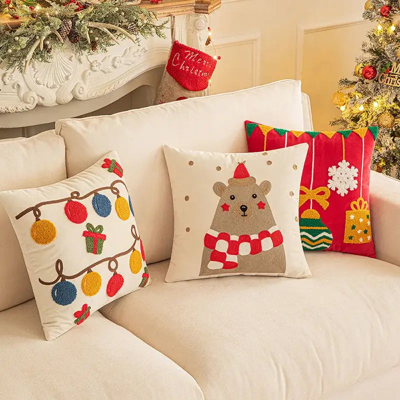 Three festive christmas embroidered red pillow covers by decorbites featuring holiday lights, bear, and gift designs on a cream sofa