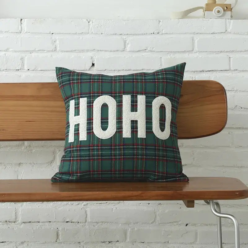 A festive green plaid decorbites christmas letter cushion cover with bold white ho ho embroidery on a wooden bench
