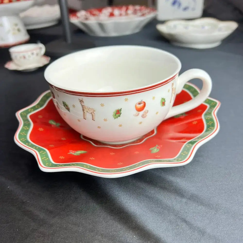 A festive white ceramic teacup with red and green holiday illustrations on a matching scalloped saucer from decorbites christmas porcelain tableware set