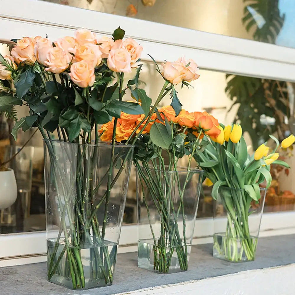 Three clear acrylic hurricane flower vases in varying sizes holding peach roses orange roses and yellow tulips
