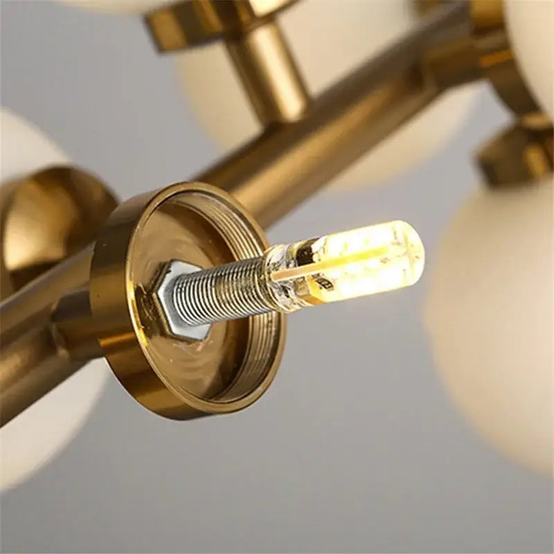 A warm glowing led bulb with clear glass and visible filament mounted in brass fixture of decorbites contemporary chandelier with white glass ball