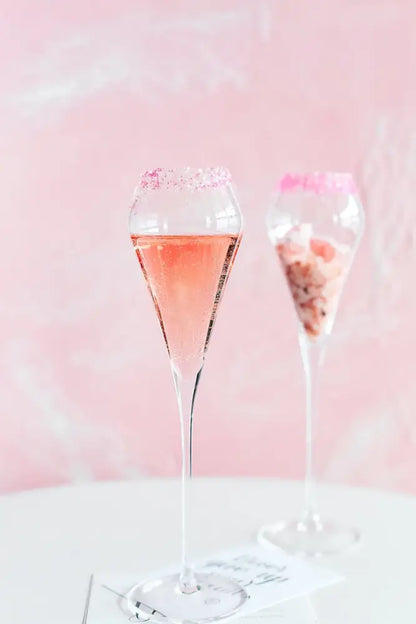 A tall slender decorbites crystal champagne flute filled with pink sparkling liquid rimmed with pink sugar crystals