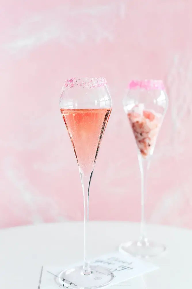A tall slender decorbites crystal champagne flute filled with pink sparkling liquid rimmed with pink sugar crystals