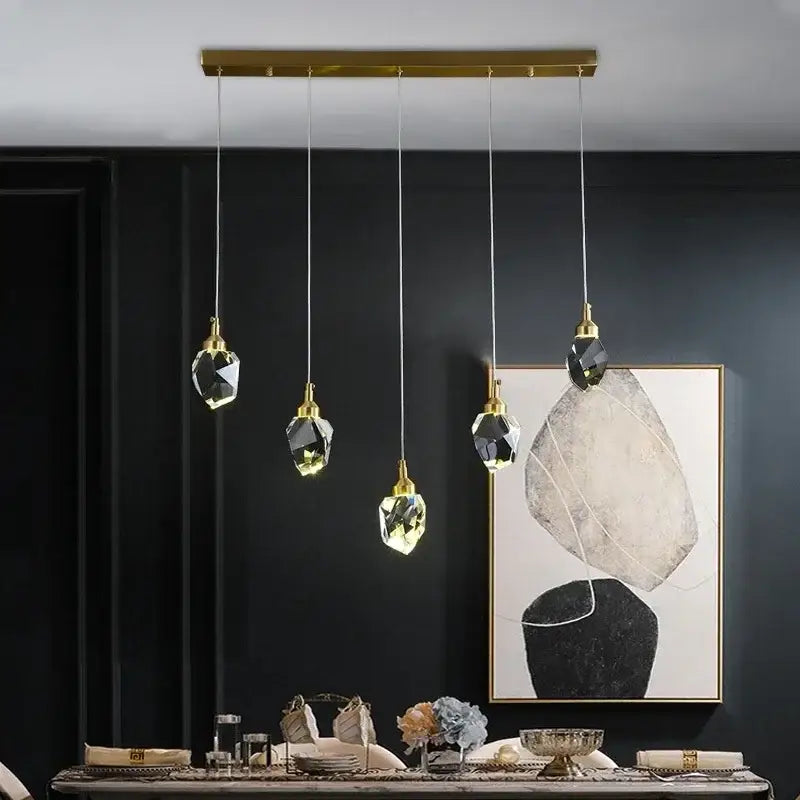 Five faceted crystal pendant lights with gold-toned fixtures hang from a brass bar in DecorBites crystal chandelier LED design