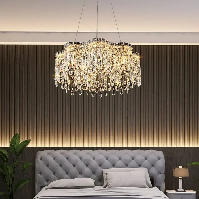 A decorbites crystal chandelier with cascading clear glass prisms and a sleek metallic frame above a tufted gray bed