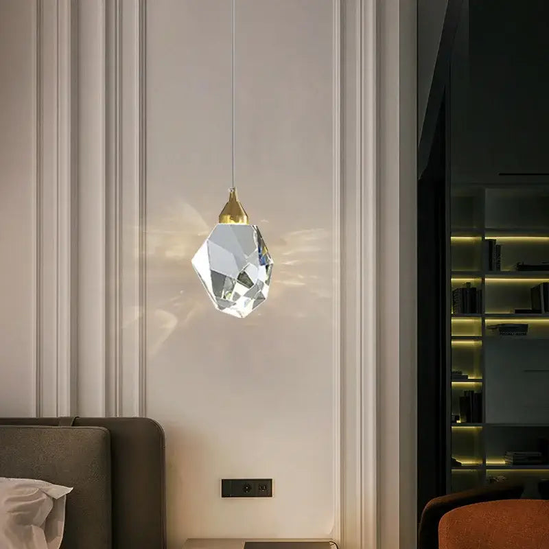 A faceted crystal chandelier pendant with gold-toned top hanging on thin cord against white paneled wall by decorbites