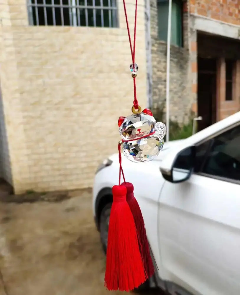 A clear faceted crystal lucky cat car hanging ornament with red tassels and matching cord for living room decor