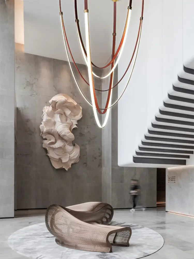 A sculptural beige-toned lounge chair with woven mesh and curved metal frame beside a flexible leather belt led chandelier