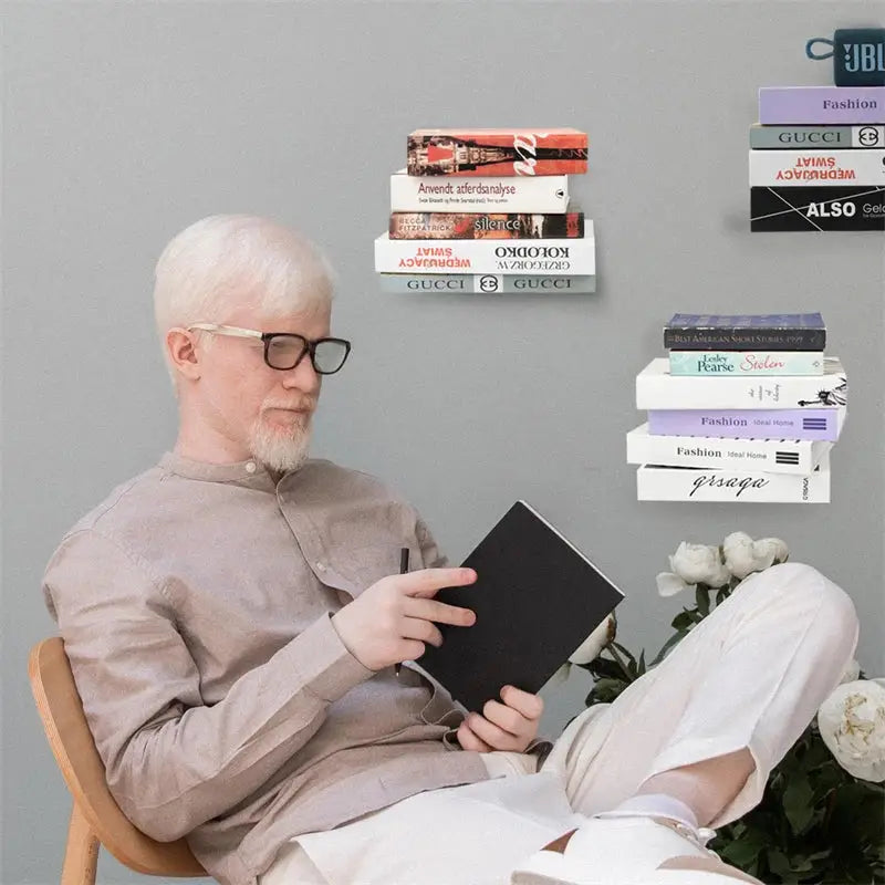 A man with white hair and glasses reads a book while sitting near beige 2pcs floating metal bookshelves by decorbites