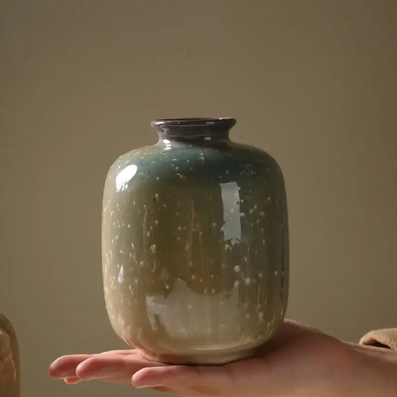 A hand holds a 13.5cm high decor ceramic vase with glossy teal to beige glaze and white speckles by DecorBites