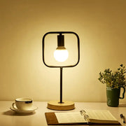 Modern black geometric frame metal table lamp with warm glow, square glass enclosure, and round wooden base for bedside use