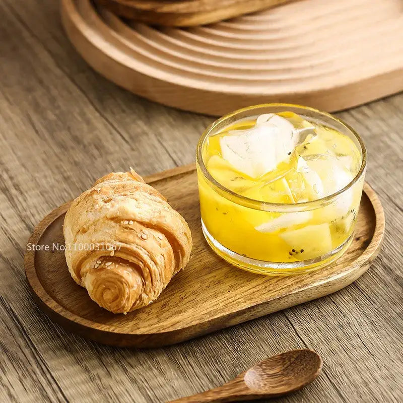A golden-brown croissant with sesame seeds beside a yellow drink on an oval wooden tray with decorbites glass afternoon tea bowl