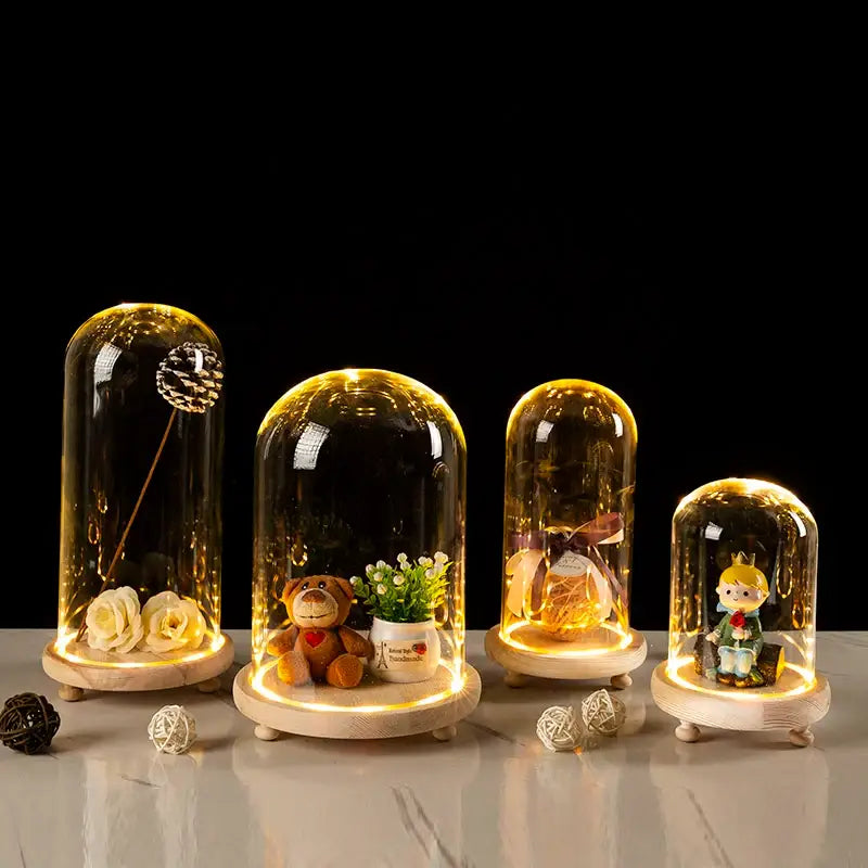 Four illuminated glass bell jar terrariums with wooden bases each displaying a unique miniature decorative item