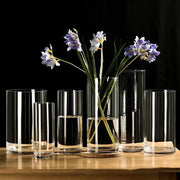 Clear cylindrical glass planter vases of varying heights with subtle vertical ridges, one holding purple flowers on wood coaster