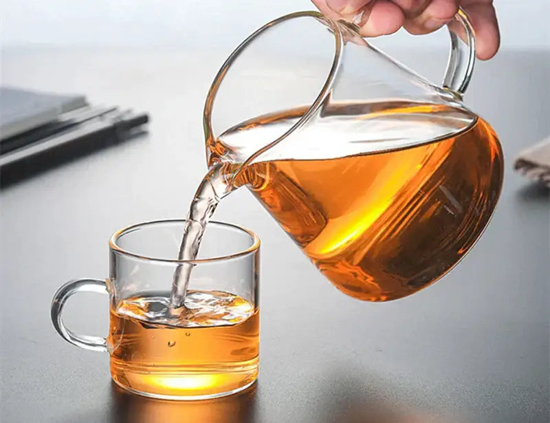 A clear glass tea pot set with a curved handle pours amber tea into a matching heat-resistant glass mug