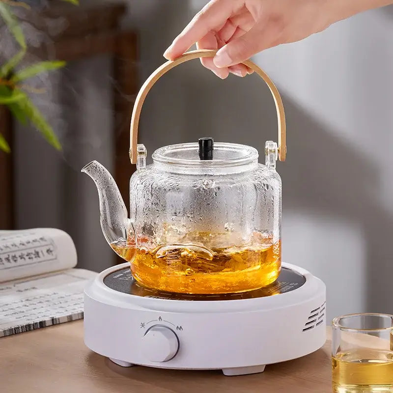Decorbites glass tea pot with a clear glass body gold handle and black lid on a white electric heating base