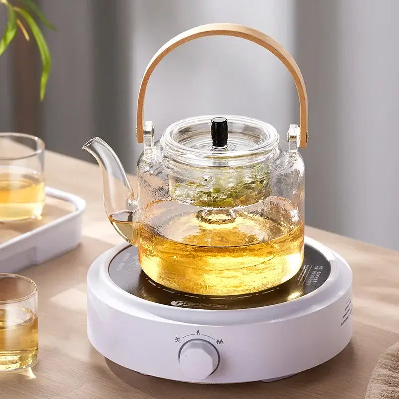 DecorBites glass tea pot with clear glass body bamboo handle and black lid knob on white electric heating base