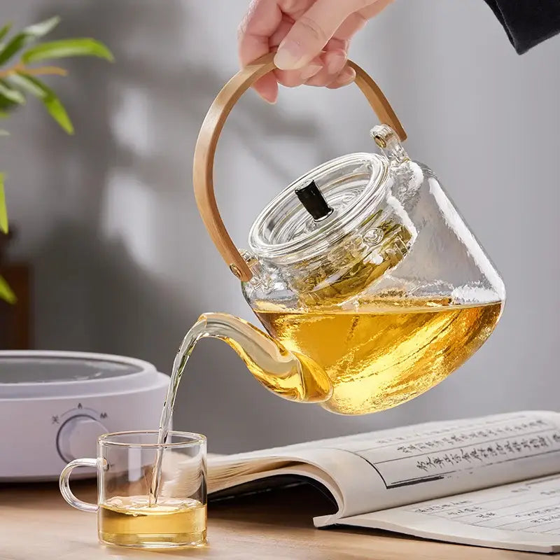 Clear glass DecorBites glass tea pot with bamboo handle pouring golden tea into a matching glass cup with double filter liner