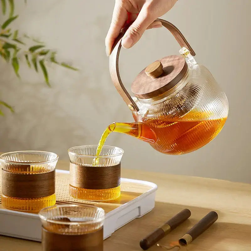 Decorbites glass tea pot with steel filter and wooden handle pouring amber tea into matching glass cups