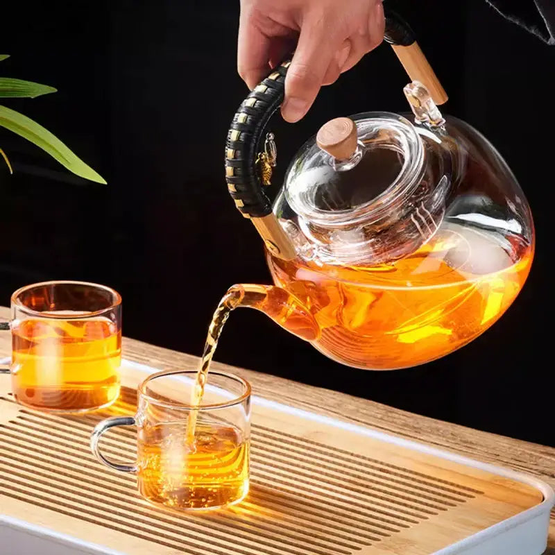Clear glass decorbites glass tea pot with wooden weave handle pouring amber tea into matching heat-resistant glasses