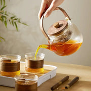 Clear glass tea pot with wooden handle and lid pouring amber tea into matching glasses decorbites glass tea set