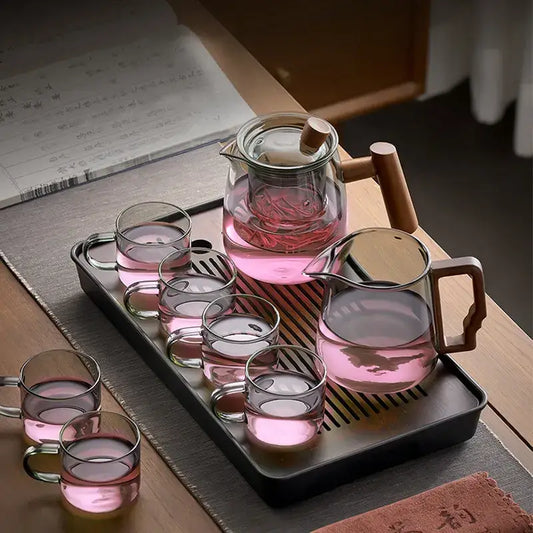 Clear glass decorbites glass tea pot with wooden handle and lid on black tray with glasses filled with pink tea