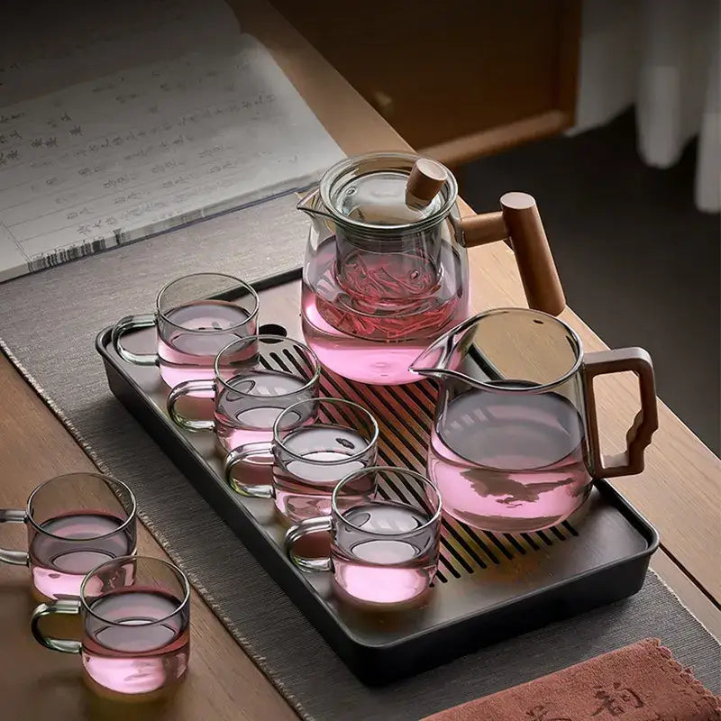 Clear glass decorbites glass tea pot with wooden handle and lid on black tray with glasses filled with pink tea