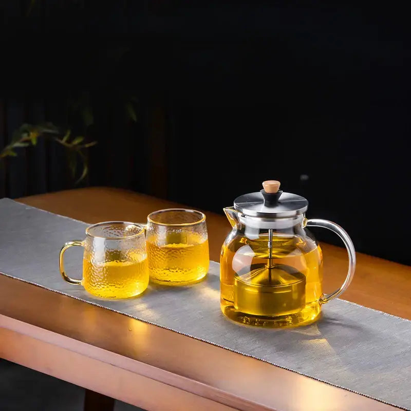 Clear glass tea set with stainless steel lid wooden knob and two textured mugs filled with golden tea by decorbites glass tea