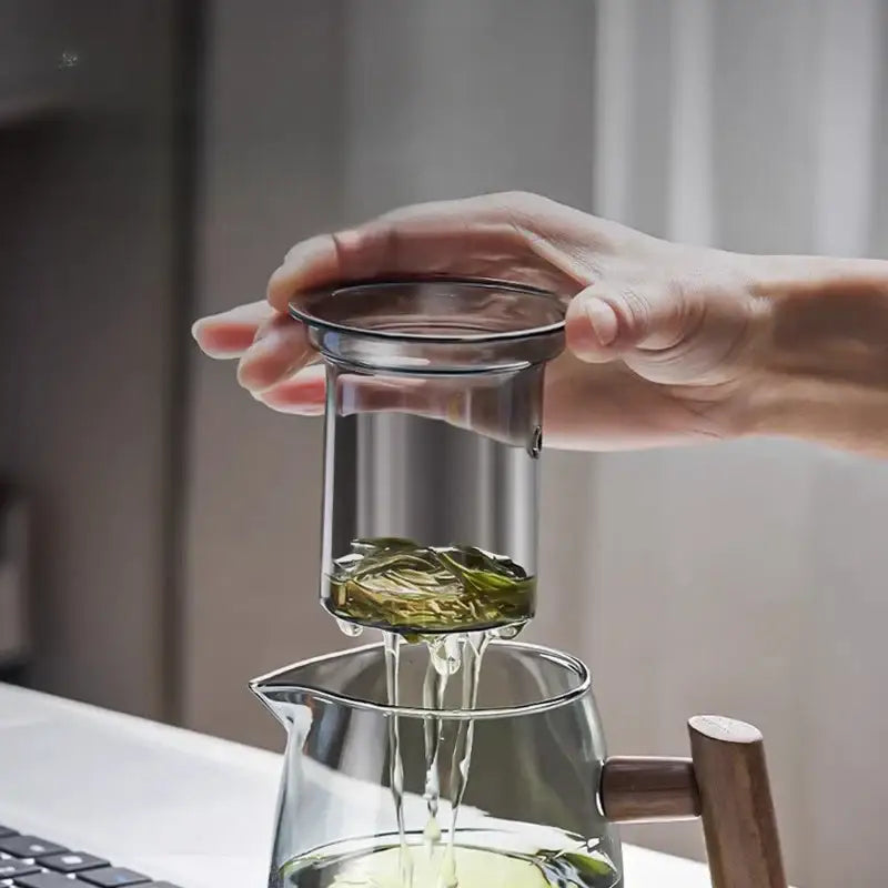 Decorbites™ glass tea set with solid wood handle and metal rim straining green leaves into clear glass teapot